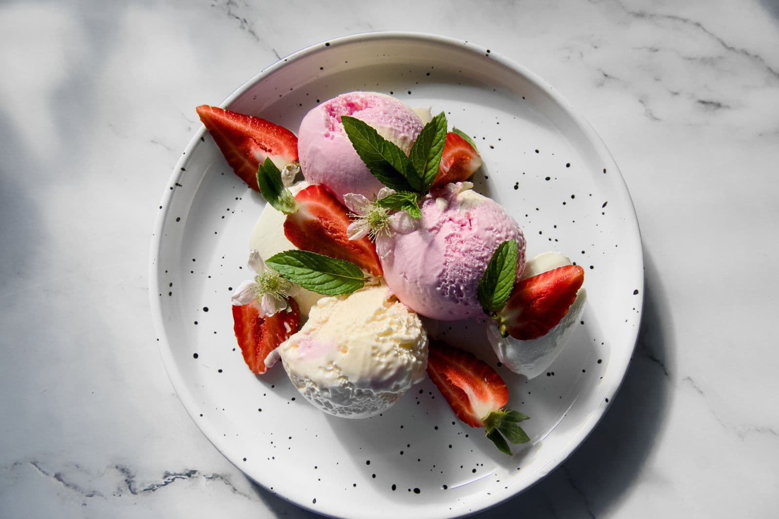 Scoops of pink strawberry and pale vanilla gelato on a speckled plate with fresh strawberries and mint