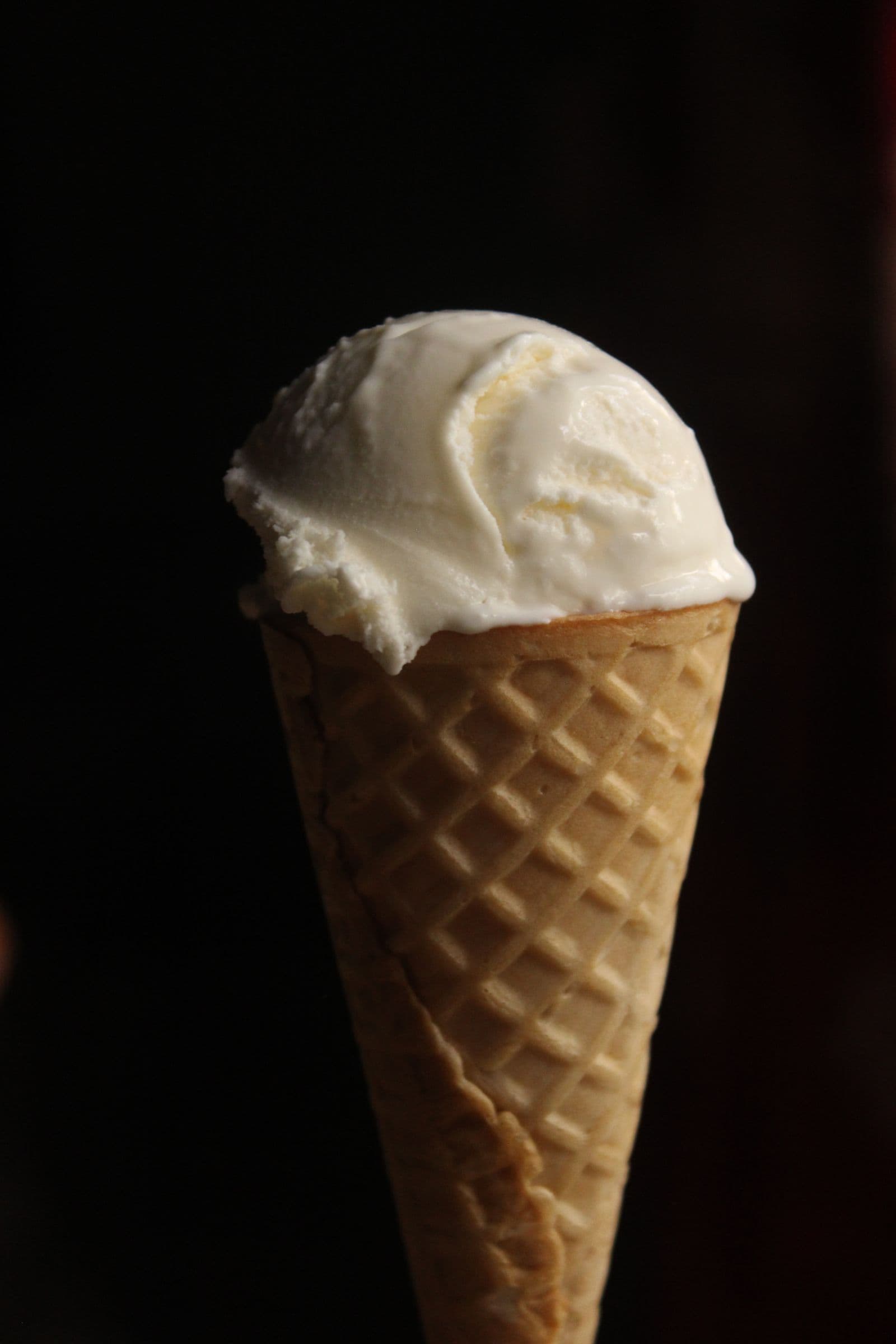 A single scoop of vanilla gelato on a waffle cone against a dark background