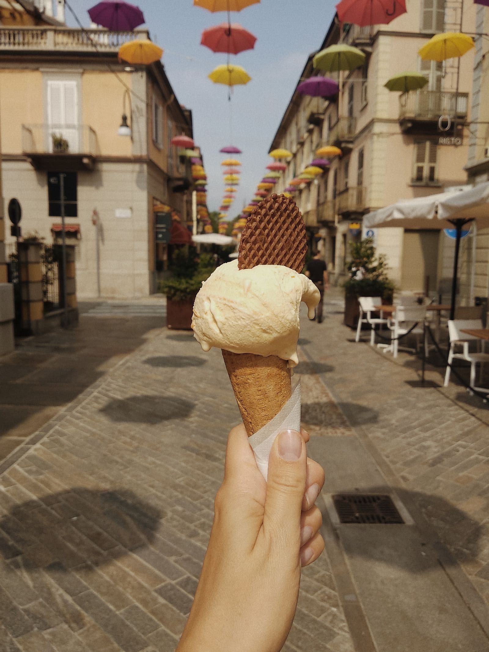 A hand holding a waffle cone of pale gelato on an Italian street strung with coloured umbrellas