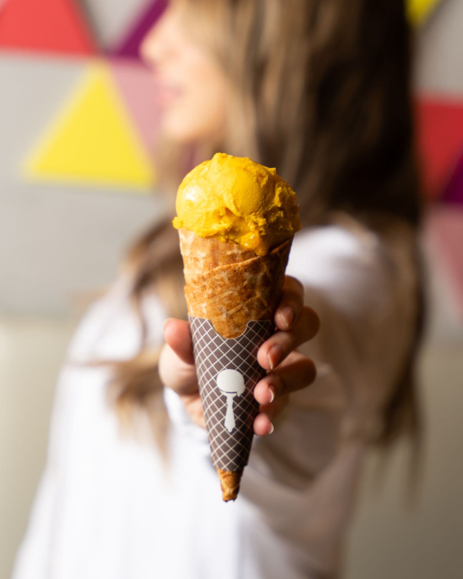 A hand holding a waffle cone topped with a deep golden scoop of peach sorbet