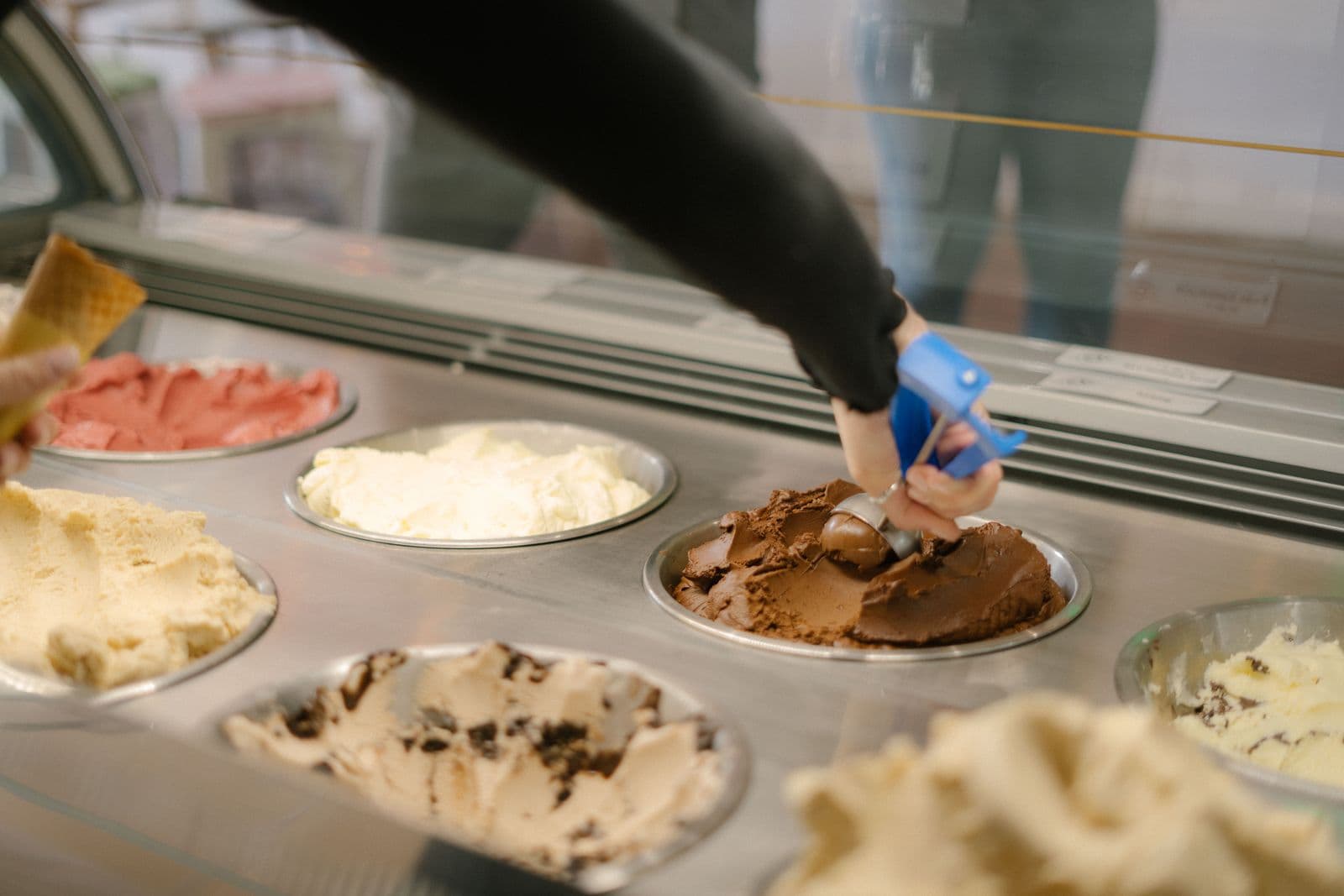A gloved hand lifting a scoop of chocolate gelato from a tray behind the counter