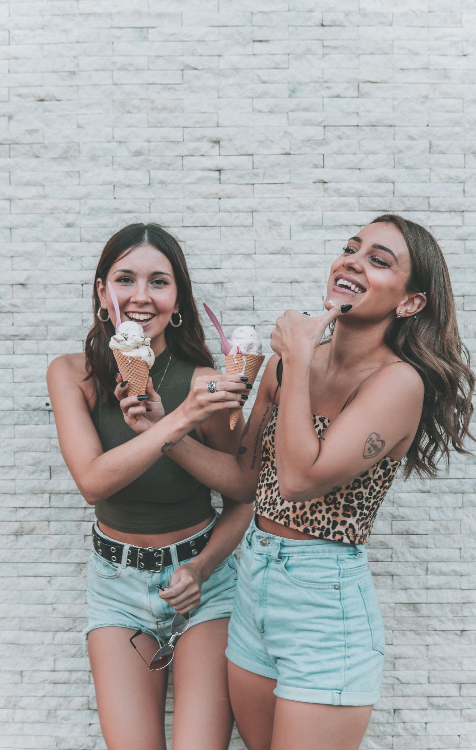 Two friends laughing together holding gelato cones against a pale brick wall
