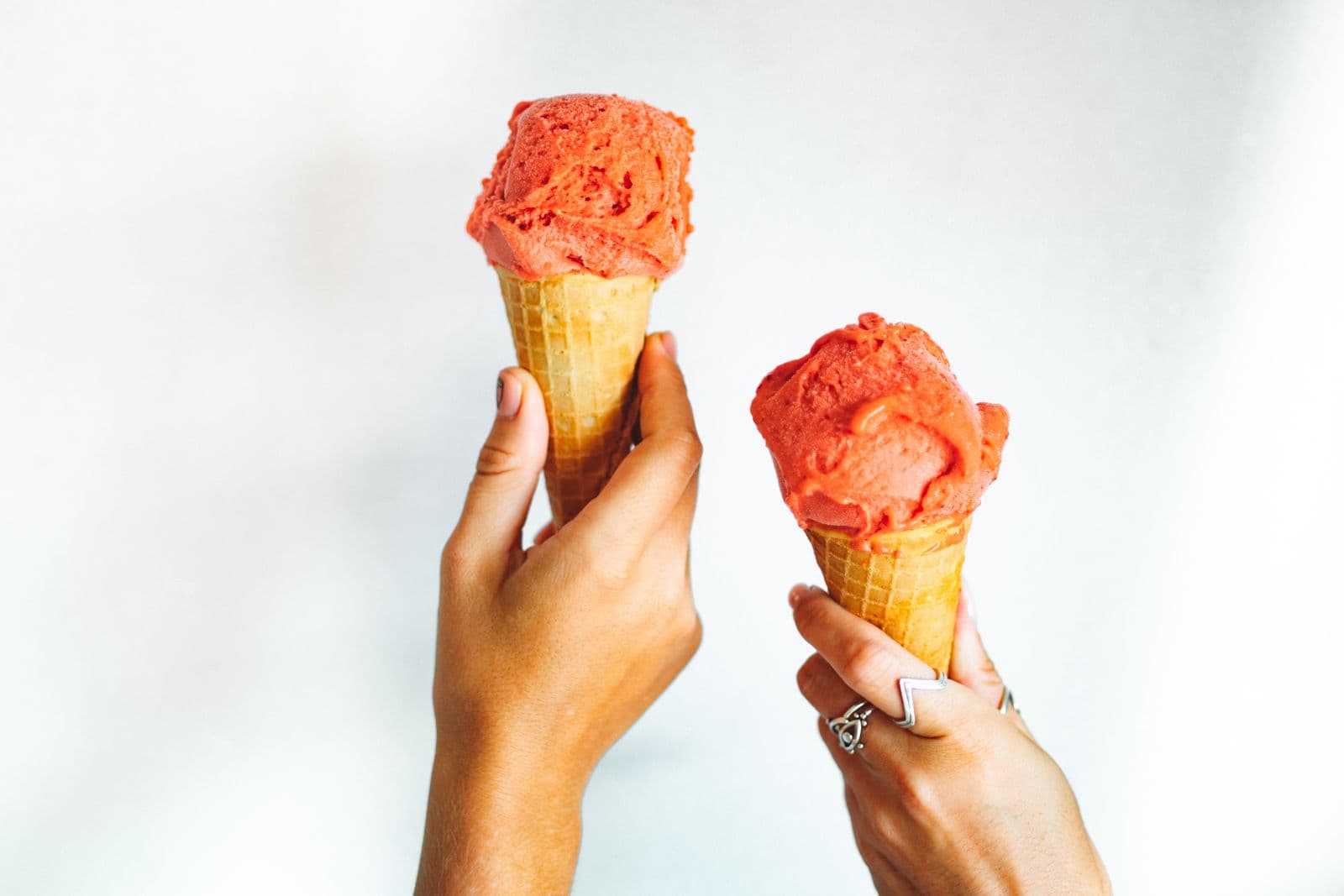 Two hands raising cones of bright red sorbet against a plain white wall
