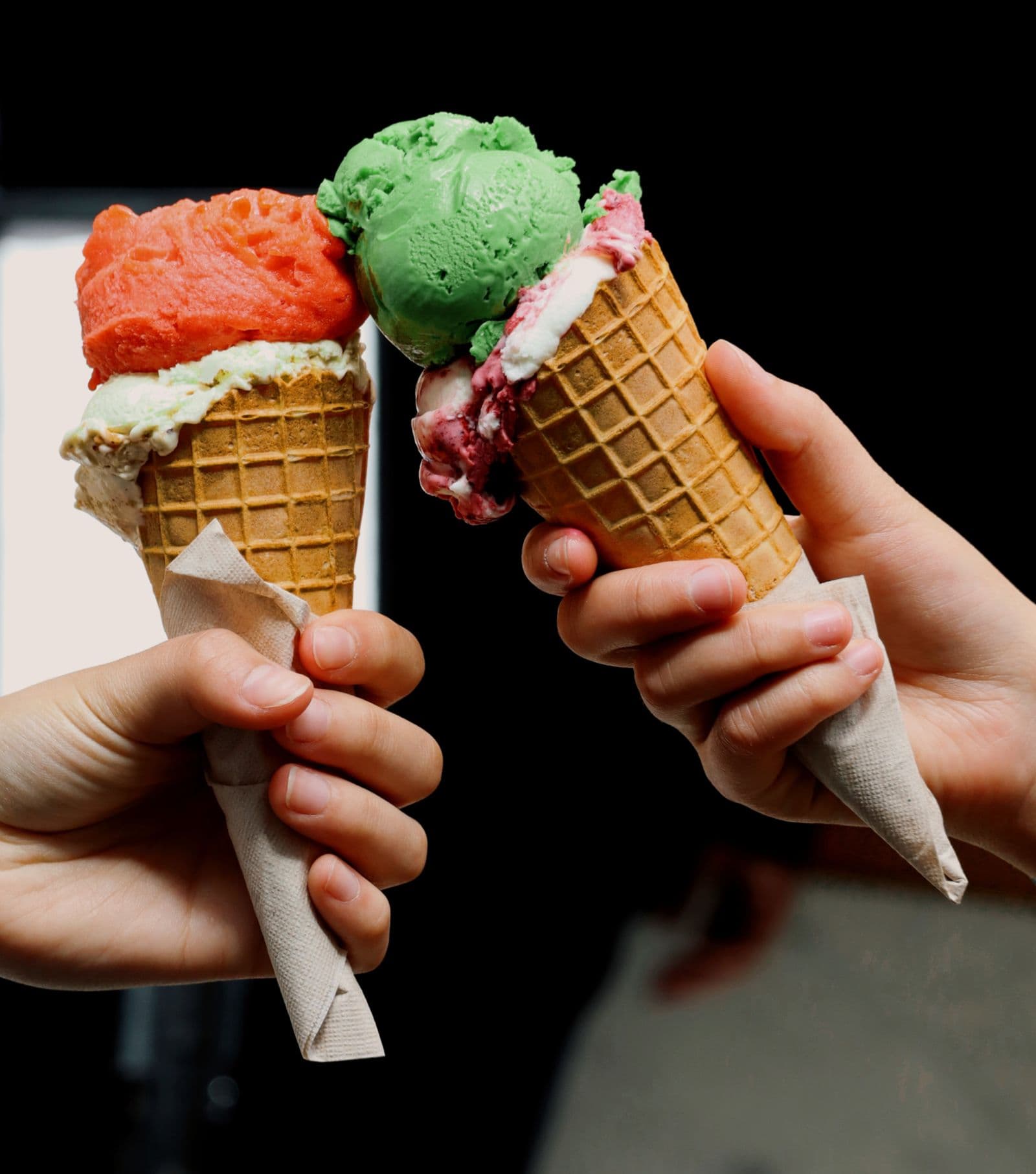 Two hands holding waffle cones of pistachio and red sorbet gelato against a dark background