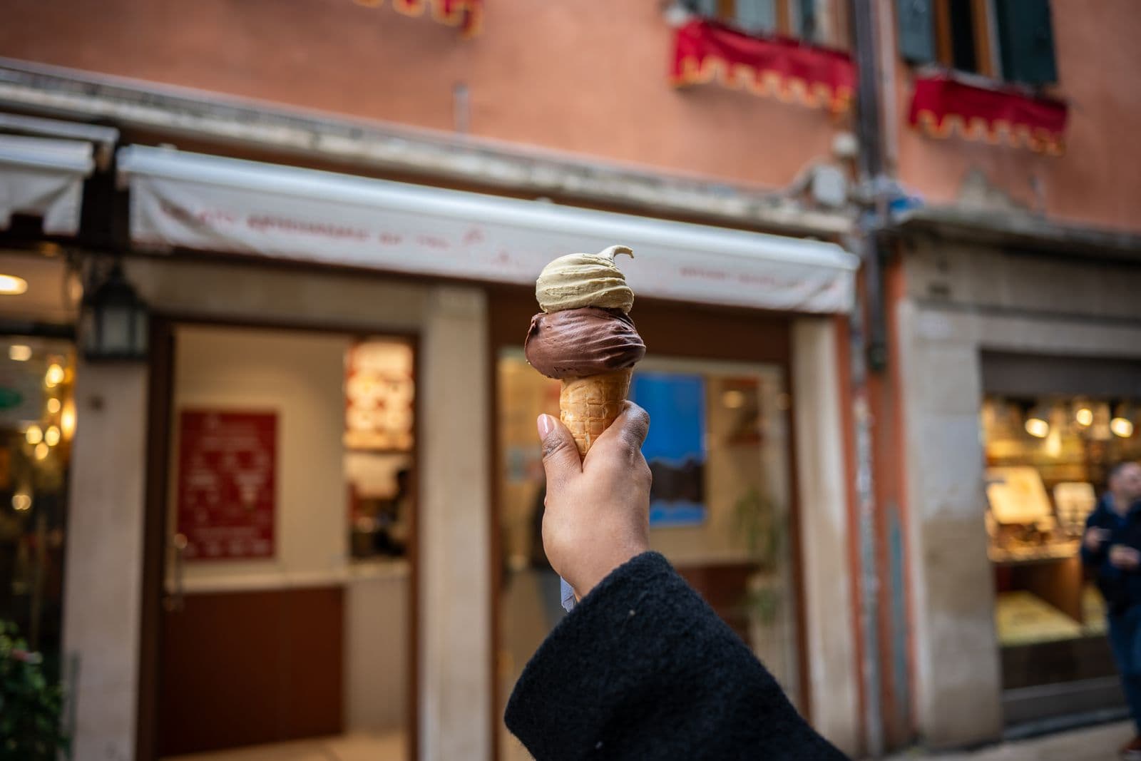 A hand holding a two-scoop gelato cone in front of a warm-toned Venetian shopfront