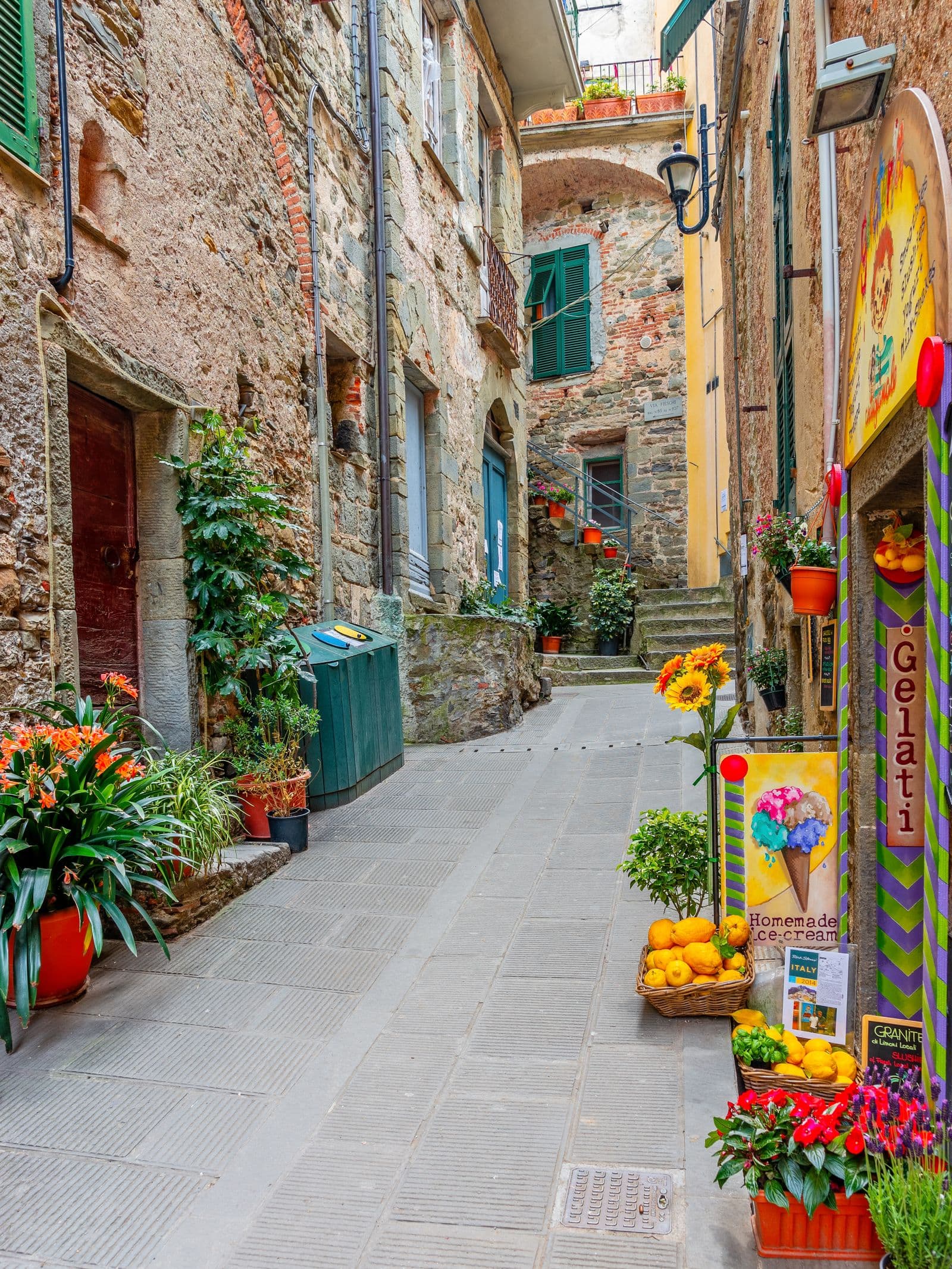 A narrow Italian alleyway of stone houses, flower pots and a gelateria sign stacked with lemons